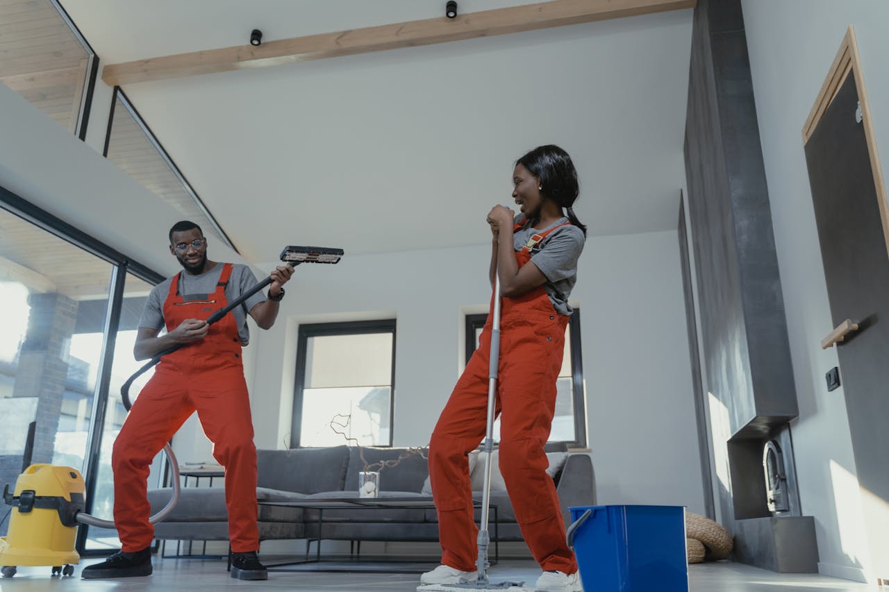services-02 Two adults in vibrant coveralls cleaning a modern home interior, enjoying work.
