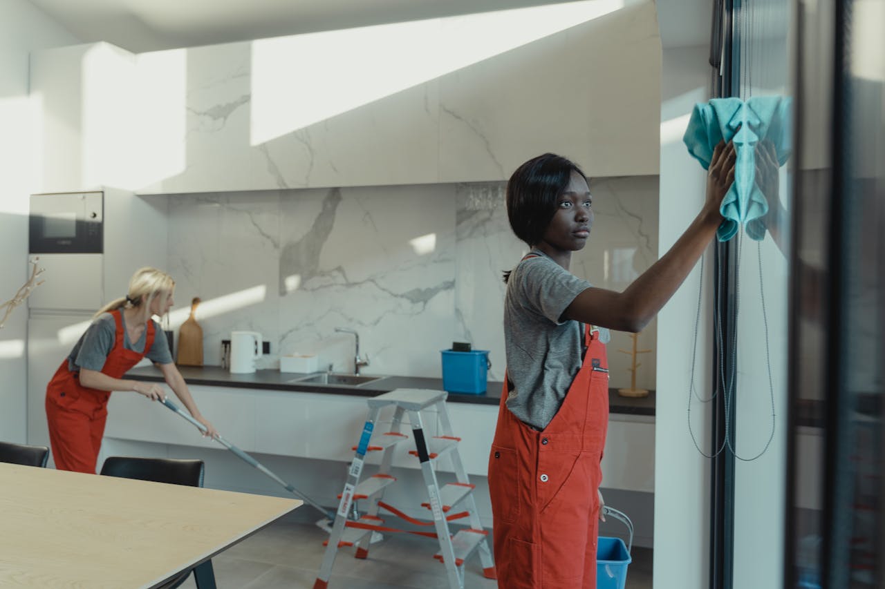 gallery-6 Two women cleaning a modern kitchen, wiping glass surfaces and mopping the floor.